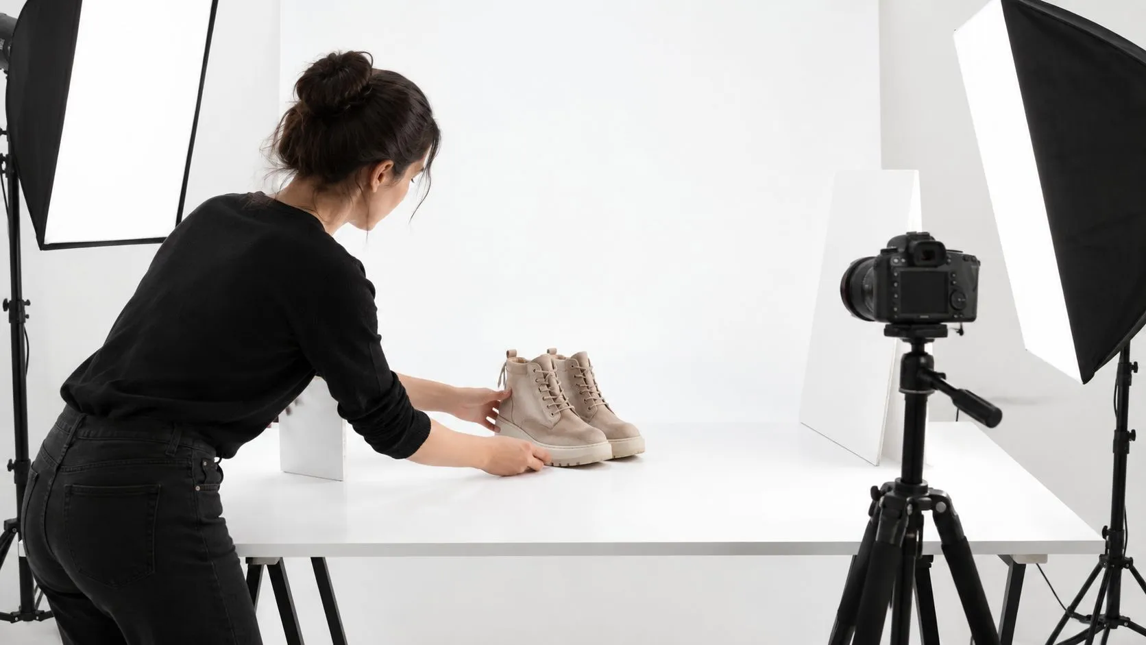Une photographe professionnelle ajuste des chaussures beige sur une table blanche dans un studio photo lumineux.